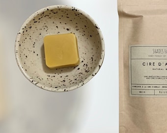 May include: A square, yellow wax melt sits in a speckled ceramic bowl. A brown paper bag with the text "CIRE D'A" and "NATURAL" is visible. The bag also has the text "SHADES FA" and "HAND POURED SOY".