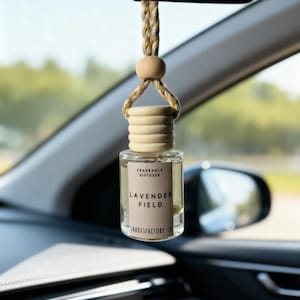 Könnte beinhalten: Ein klarer Auto-Lufterfrischer aus Glas mit einem Holzaufsatz und einem beigen Etikett mit der Aufschrift "Lavender Field". Der Lufterfrischer hängt am Rückspiegel eines Autos. Der Text "Fragrance Diffuser" ist ebenfalls sichtbar.