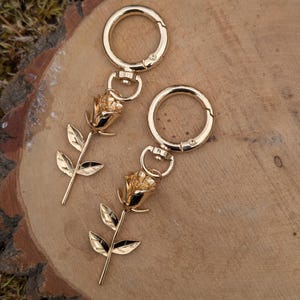 May include: Two gold-colored keychains, each featuring a detailed rose design. The roses have intricate petals and are attached to a clasp. The keychains are displayed on a natural wood surface, creating a rustic aesthetic.