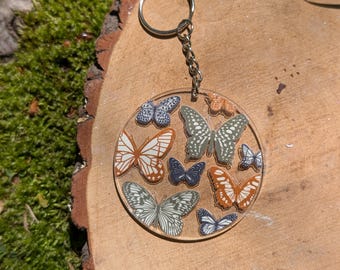 May include: A round, clear keychain with a silver key ring. The keychain features a design of multiple butterflies in various shades of brown, green, and black.
