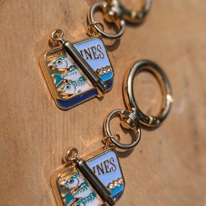 May include: Two gold-colored keychains with a sardine can design. Each keychain features a rectangular charm with the word "INES" on a blue background, a sardine can with three fish, and a gold-colored ring.