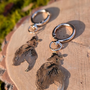 May include: Two gold-colored keychains with fish-shaped pendants. The pendants have intricate details, with a dark brown and gold color scheme. The keychains are attached to large, round, gold-colored clasps. The background is a wooden surface.