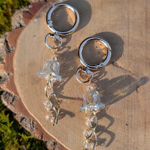 Könnte beinhalten: Zwei Schlüsselanhänger mit goldfarbenen Verschlüssen und transparenten Quallen-Anhängern. Die Quallen-Anhänger haben schillernde Oberteile, goldfarbenen Draht und klare Perlenakzente. Die Schlüsselanhänger werden auf einer Holzoberfläche präsentiert.