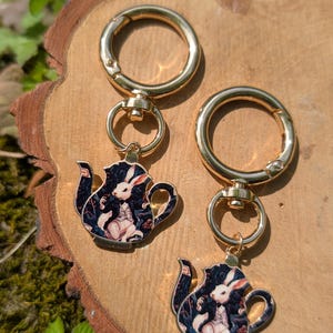 May include: Two gold-colored keychains with a teapot charm featuring a white rabbit in a waistcoat. The teapot charm has a dark blue background with floral accents. The keychains are on a wooden surface.