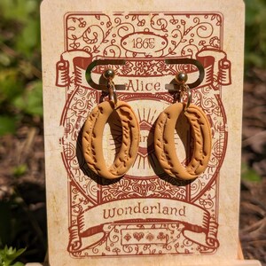 May include: A pair of brown clay earrings with a gold-tone hook closure. The earrings are shaped like an oval with a textured design. The earrings are displayed on a card with the text "Alice in Wonderland" and the year "1865".