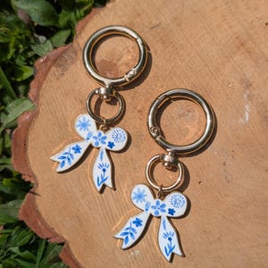 May include: Two gold keyrings, each with a white bow-shaped charm. The charms feature blue floral designs. The keyrings are displayed on a wooden surface, with green foliage visible in the background.
