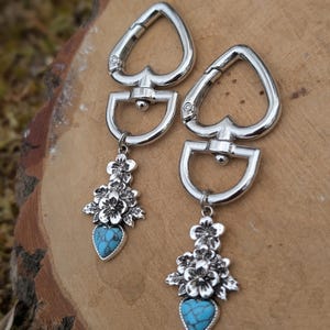 May include: A pair of silver-toned earrings. Each earring features a spade-shaped top, a D-shaped loop, and a floral cluster with a turquoise heart-shaped stone pendant. The earrings are displayed on a wooden surface.