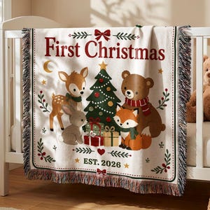 May include: A cream-colored woven throw blanket featuring the text First Christmas and EST. 2026. The design includes a central Christmas tree surrounded by a deer, a rabbit, a bear, and a fox, all accented with red and green holiday gift boxes.