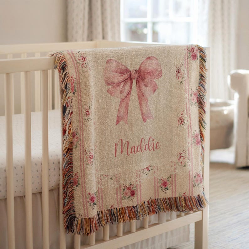 May include: A beige baby blanket featuring a large pink bow and the name "Maddie" in cursive. The blanket has a floral border and fringe trim. The blanket is draped over a white cot.