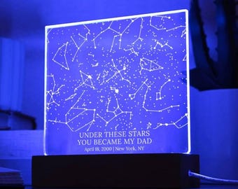 May include: A blue-lit, square-shaped night light with a constellation design. The text "UNDER THESE STARS YOU BECAME MY DAD April 18, 2000 | New York, NY" is displayed on the light. The light sits on a dark wooden base.