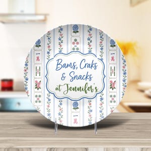 Puede incluir: Un plato redondo de cer&aacute;mica con fondo blanco, rayas verticales azules, enredaderas florales y motivos de fichas de Mahjong. El centro muestra un marco azul festoneado con el texto Bams, Craks & Snacks at Jennifer's.