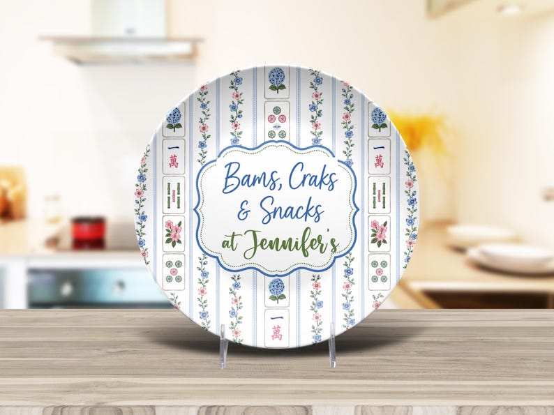 Puede incluir: Un plato redondo de cer&aacute;mica con fondo blanco, rayas verticales azules, enredaderas florales y motivos de fichas de Mahjong. El centro muestra un marco azul festoneado con el texto Bams, Craks & Snacks at Jennifer's.