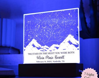 Op de afbeelding: Een gepersonaliseerd nachtlampje met een sterrenkaartontwerp. Het acryl paneel toont een sterrenbeeldkaart boven een bergketen, met de tekst "THE STARS ON THE NIGHT YOU WERE BOTN" en een naam, datum en locatie. De basis is zwart en biedt 7 lichtkleuren.