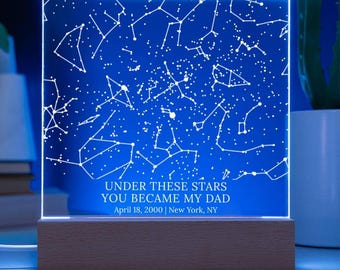 May include: A blue LED night light with a constellation design. The light has white star patterns and the text "UNDER THESE STARS YOU BECAME MY DAD April 18, 2000 | New York, NY". The light sits on a wooden base.