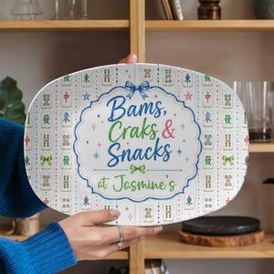 Puede incluir: Una bandeja de servir ovalada de color blanco con un patr&oacute;n repetitivo de fichas de mahjong de colores. El centro muestra un marco festoneado azul con un lazo, que contiene el texto Bams, Craks & Snacks at Jasmine's en azul, verde y rosa.