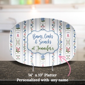 Puede incluir: Una bandeja ovalada de 35,56 cm por 25,4 cm con fondo blanco, rayas verticales azules, enredaderas florales e ilustraciones de fichas de mahjong. El centro muestra un marco festoneado azul con el texto Bams, Craks & Snacks at Jennifer's.