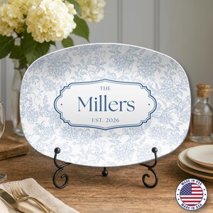 Puede incluir: Fuente ovalada con diseño floral azul y el texto "The Millers EST. 2026". La fuente se exhibe sobre un soporte de metal negro. La fuente tiene un fondo blanco con patrones florales azules.