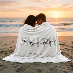 May include: A white fleece blanket featuring the large grey text SMITH, with the names henry and emily written in black cursive script separated by a small heart, and the text Est. 10.22.2024 printed in black below the names.
