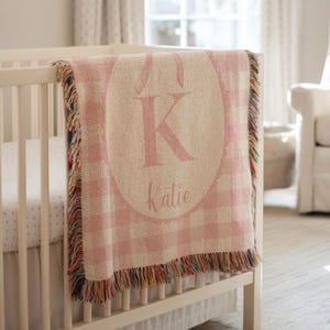 May include: A pink and white checked baby blanket with a large pink letter "K" and the name "Katie" embroidered on it. The blanket has a fringed edge and is draped over a white cot.