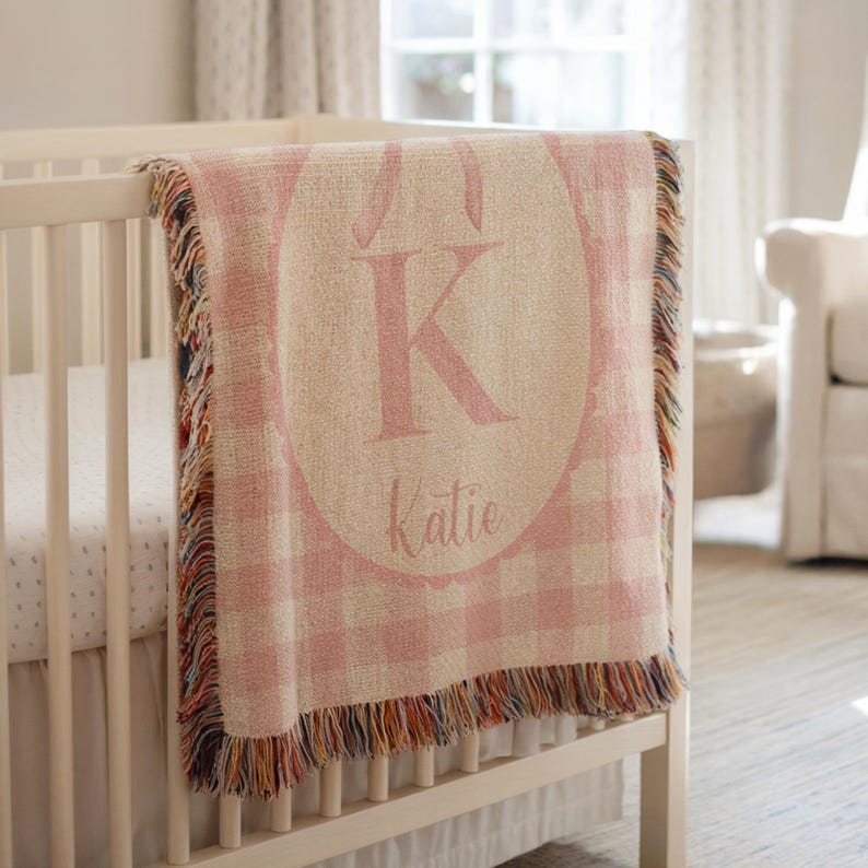 May include: A pink and white checked baby blanket with a large pink letter "K" and the name "Katie" embroidered on it. The blanket has a fringed edge and is draped over a white cot.