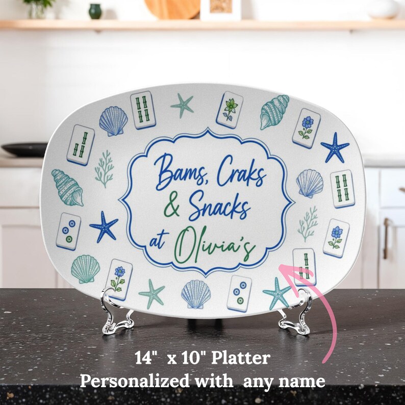 Puede incluir: Una bandeja ovalada blanca de 35,5 cm por 25,4 cm con un patr&oacute;n azul y verde de conchas, estrellas de mar y fichas de mahjong. El centro muestra el texto Bams, Craks & Snacks at Olivia's dentro de un borde azul festoneado.