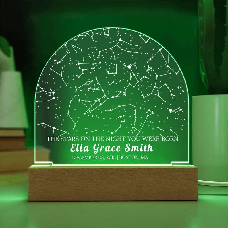 May include: A personalized night light with a constellation design. The green-lit acrylic panel displays a star map and the text "THE STARS ON THE NIGHT YOU WERE BORN" with a name and date. It sits on a wooden base.