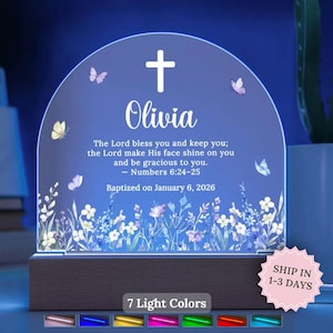 May include: A personalized baptism night light with a cross and the name "Olivia" on a blue background. The light includes a Bible verse and butterfly accents. The base is dark wood, and it offers 7 light colors.