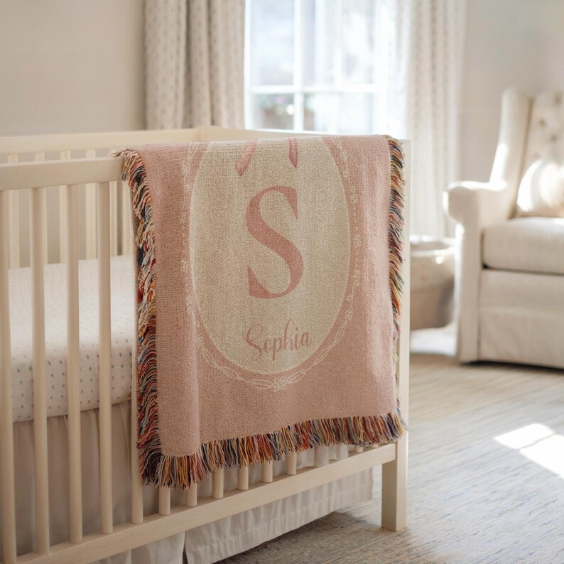 May include: A pink woven baby blanket with a cream oval design, featuring the letter "S" and the name "Sophia" in pink. The blanket has a fringed edge with multiple colours and is draped over a white cot.