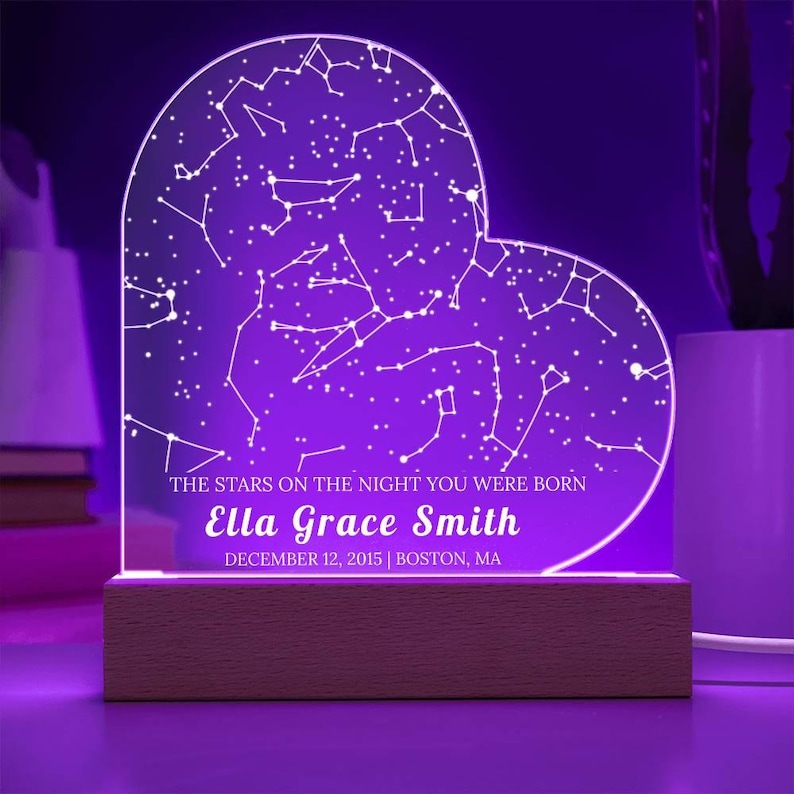 May include: A heart-shaped night light with a constellation design, illuminated in purple. The base is made of wood. Text on the light reads "THE STARS ON THE NIGHT YOU WERE BORN" and "Ella Grace Smith, DECEMBER 12, 2015 | BOSTON, MA."