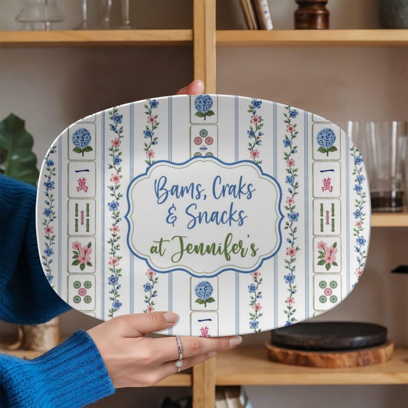 Puede incluir: Una bandeja de servir ovalada con fondo blanco, rayas azules verticales, enredaderas florales y motivos de fichas de mahjong. El centro muestra un marco azul decorativo con el texto "Bams, Craks & Snacks at Jennifer's" en letras azules y verdes.