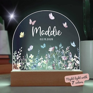 Könnte beinhalten: Ein weißes Acryl-Nachtlicht in Halbkreisform mit einem Blumendesign und Schmetterlingen. Das Licht trägt den Namen "Maddie" und das Datum "02.15.2025". Der Sockel des Lichts ist aus Holz und der Text "Nachtlicht mit 7 Farben" ist in der rechten unteren Ecke sichtbar.