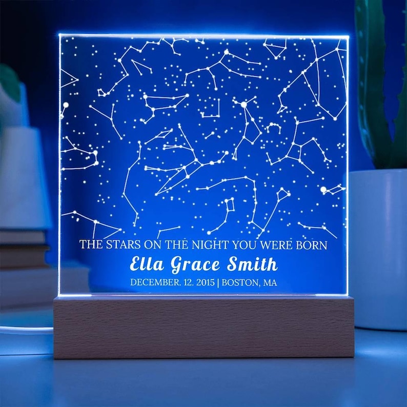 May include: A personalized night light with a blue background displaying a constellation map. The text reads "THE STARS ON THE NIGHT YOU WERE BORN" and "Ella Grace Smith, December 12, 2015 | Boston, MA." The light sits on a wooden base.