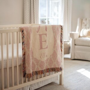May include: A pink and cream-coloured baby blanket with the letter "E" and the name "Ella" displayed on a cot. The blanket has a decorative pattern and fringe trim. The cot is white.