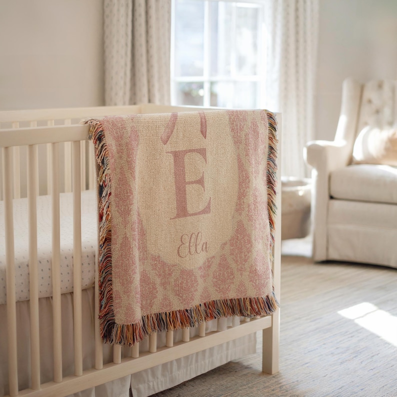 May include: A pink and cream-coloured baby blanket with the letter "E" and the name "Ella" displayed on a cot. The blanket has a decorative pattern and fringe trim. The cot is white.