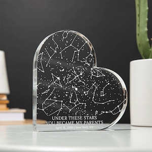 May include: A clear, heart-shaped acrylic keepsake featuring a constellation design. The text "UNDER THESE STARS YOU BECAME MY PARENTS" is inscribed, along with the date April 18, 2000, and New York, NY. The keepsake is displayed on a white surface.