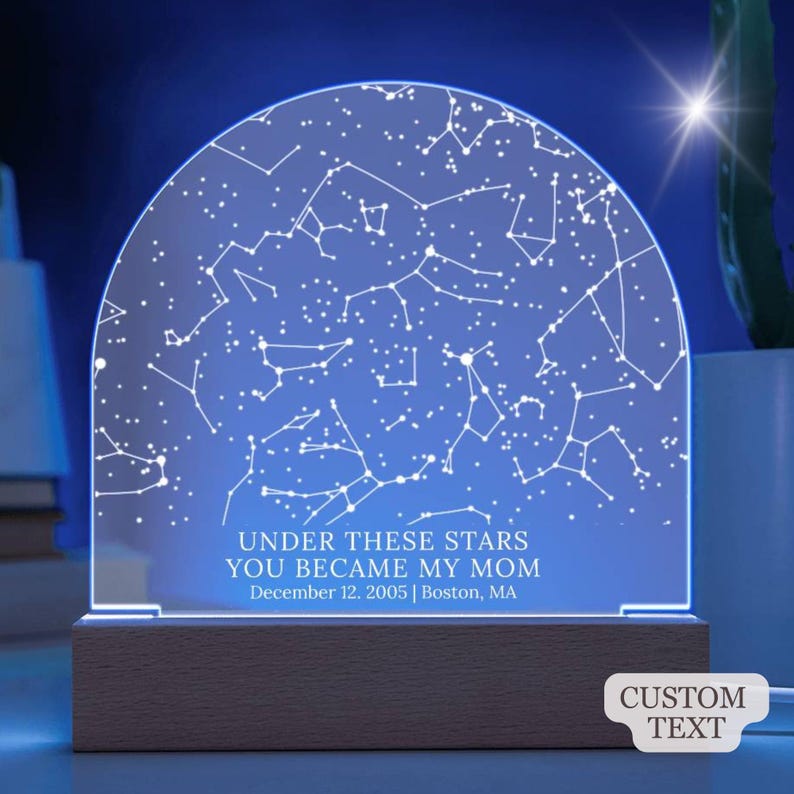 May include: A personalized night light featuring a constellation map design. The acrylic panel displays a blue starry sky with connected white dots and lines. Text reads "UNDER THESE STARS YOU BECAME MY MOM" with a date and location. The base is made of wood.