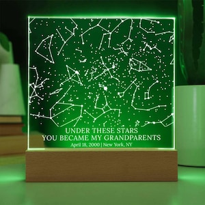 May include: A green illuminated night light with a constellation design. The text "UNDER THESE STARS YOU BECAME MY GRANDPARENTS April 18, 2000 | New York, NY" is displayed. The light sits on a wooden base.