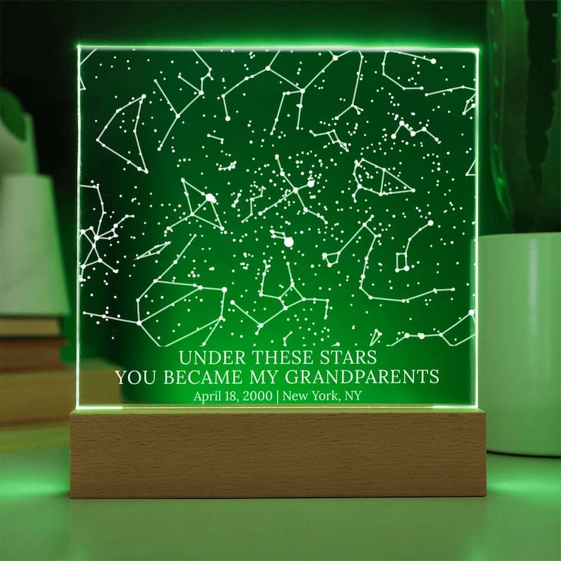May include: A green illuminated night light with a constellation design. The text "UNDER THESE STARS YOU BECAME MY GRANDPARENTS April 18, 2000 | New York, NY" is displayed. The light sits on a wooden base.