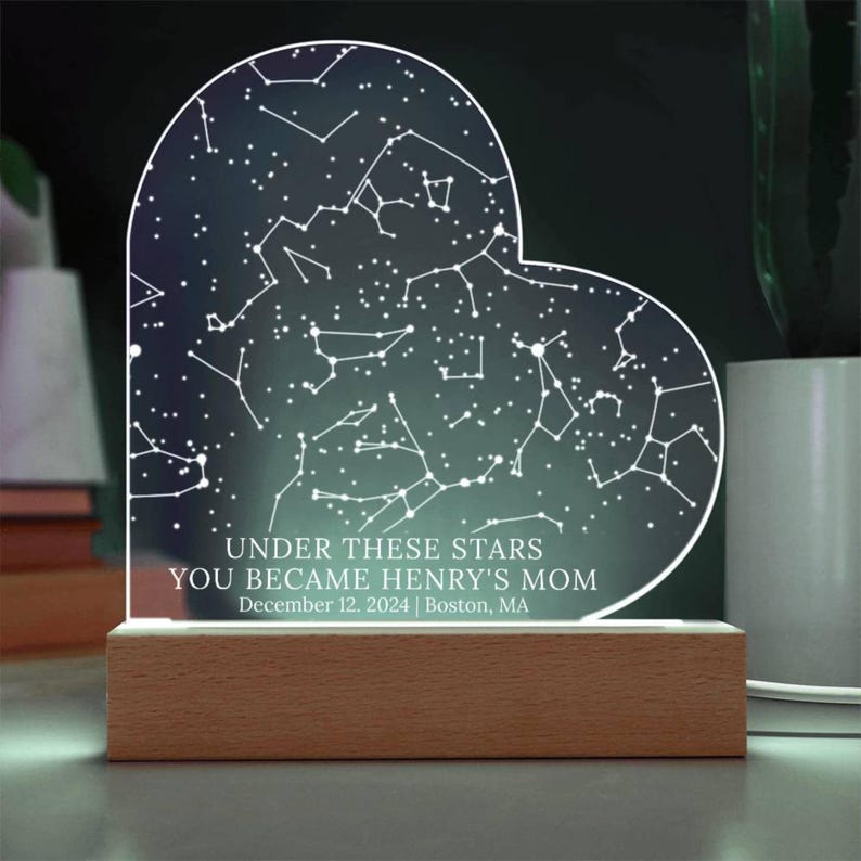 May include: Heart-shaped night light featuring a constellation design with the text "UNDER THESE STARS YOU BECAME HENRY'S MOM December 12, 2024 | Boston, MA." The light sits on a rectangular wooden base. The background is dark.