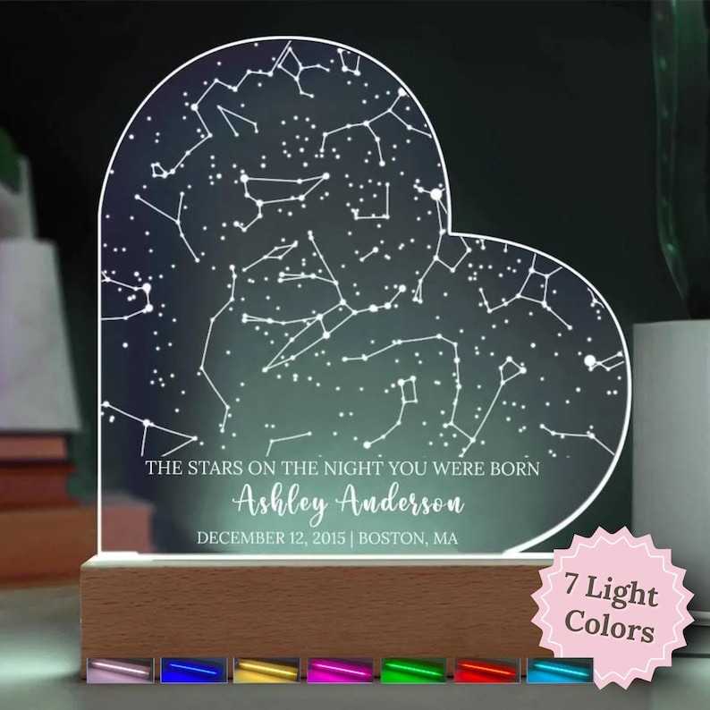 May include: A heart-shaped night light with a constellation design. The text reads "THE STARS ON THE NIGHT YOU WERE BORN" with a name and date. The base is wood, and it offers 7 light colors.