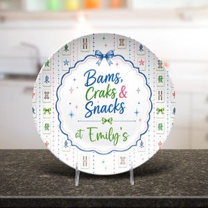 Puede incluir: Un plato redondo de cer&aacute;mica blanca con un borde decorativo de fichas de mahjong coloridas y peque&ntilde;as estrellas. El centro muestra el texto azul y verde "Bams, Craks & Snacks at Emily's" enmarcado por una l&iacute;nea azul festoneada y un lazo.