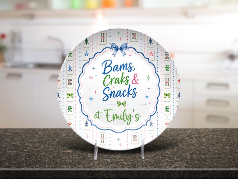 Puede incluir: Un plato redondo de cer&aacute;mica blanca con un borde decorativo de fichas de mahjong coloridas y peque&ntilde;as estrellas. El centro muestra el texto azul y verde "Bams, Craks & Snacks at Emily's" enmarcado por una l&iacute;nea azul festoneada y un lazo.