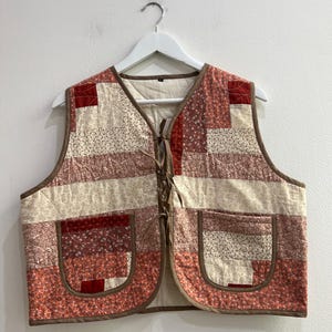 May include: A sleeveless vest featuring a patchwork design with various floral and geometric patterns in shades of red, pink, and cream. The vest has a V-neckline, two front patch pockets, and a front tie closure with brown fabric trim.