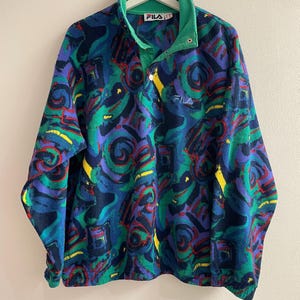 K&ouml;nnte beinhalten: Ein lang&auml;rmeliger Fleece-Pullover mit einem lebendigen abstrakten Muster in Blau-, Gr&uuml;n-, Lila- und Gelbt&ouml;nen. Das Kleidungsst&uuml;ck hat einen hohen Kragen mit Druckknopfleiste, ein gr&uuml;nes Innenfutter und das Fila-Logo auf der Brust und am Etikett.