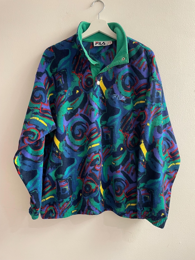 K&ouml;nnte beinhalten: Ein lang&auml;rmeliger Fleece-Pullover mit einem lebendigen abstrakten Muster in Blau-, Gr&uuml;n-, Lila- und Gelbt&ouml;nen. Das Kleidungsst&uuml;ck hat einen hohen Kragen mit Druckknopfleiste, ein gr&uuml;nes Innenfutter und das Fila-Logo auf der Brust und am Etikett.