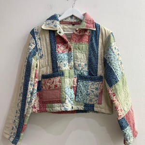 Peut inclure: Une veste courte matelassée présentant un motif patchwork composé de divers imprimés floraux et vichy dans des tons rouge, bleu, vert et crème. La veste possède un col, des manches longues, deux poches plaquées à l'avant et des boutons-pression dorés.