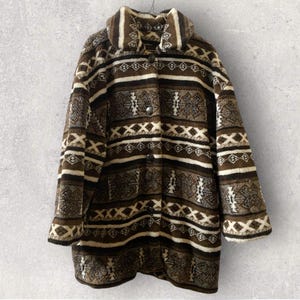 Könnte beinhalten: Ein brauner und cremefarbener Fleece-Mantel mit Muster, Knopfleiste und breitem Kragen. Der Mantel hat geometrische und gestreifte Designs in Braun-, Creme- und Schwarztönen. Der Mantel hängt vor einem grauen Hintergrund.