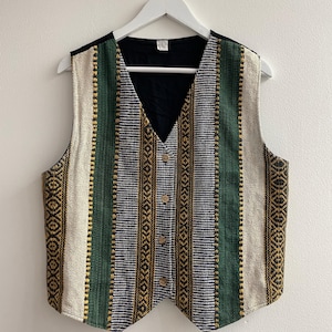 Puede incluir: Un chaleco sin mangas con un patrón tejido de rayas verticales en verde, crema y azul oscuro con detalles dorados. La prenda tiene un cuello en V, un forro interior negro y cuatro botones de madera de color claro en la parte central.