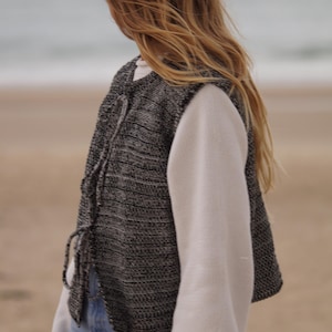 May include: A sleeveless vest with a black and white speckled pattern, tied with a drawstring, worn over a white long-sleeved shirt. The person is wearing light blue jeans. The background is a beach with the ocean in the distance.