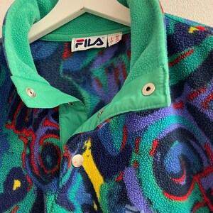 K&ouml;nnte beinhalten: Ein Fila Fleece-Pullover mit einem lebendigen, abstrakten Muster in Blau-, Gr&uuml;n-, Lila- und Gelbt&ouml;nen. Das Kleidungsst&uuml;ck verf&uuml;gt &uuml;ber einen hohen Kragen, eine kurze Knopfleiste mit silberfarbenen Druckkn&ouml;pfen und das Fila-Logo auf der Vorderseite.
